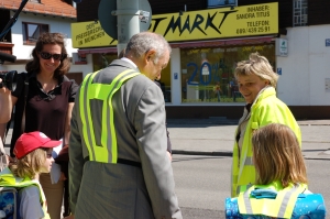 Die Schulweghelferin hilft dem Minister und den Kindern beim Überqueren der Straße am ampelgeregelten Fußgängerüberweg.